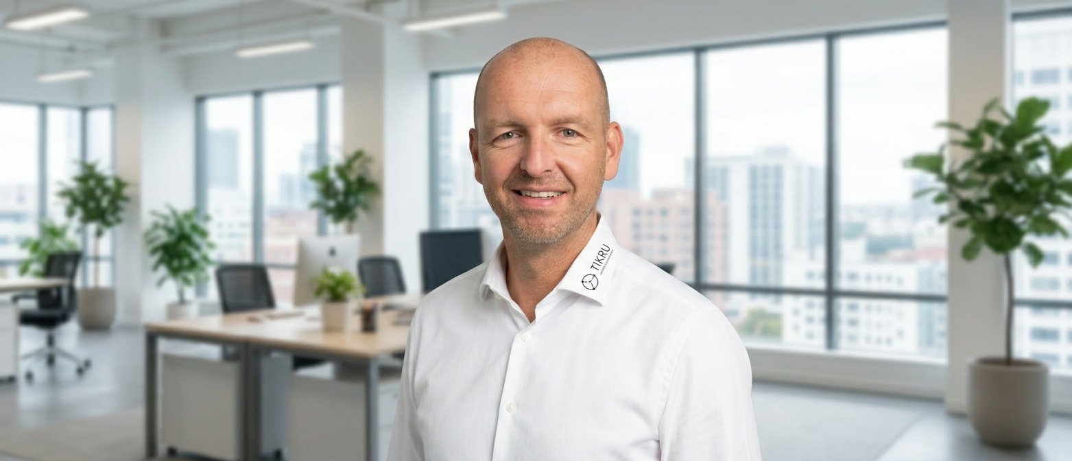 Thilo Krumm, Inhaber von TIKRU, lächelt in einem modernen Büro mit Stadtblick.